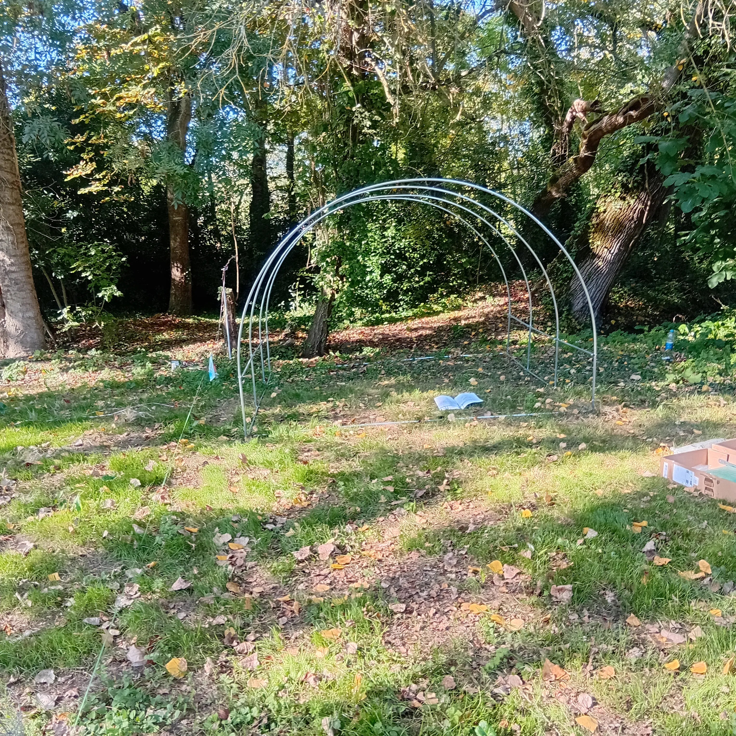 Un autre angle de la structure métallique tubulaire de la mini-serre. Les arches sont partiellement assemblées. Des cartons d'emballage ouverts sont visibles à l'arrière-plan, près des arbres Image