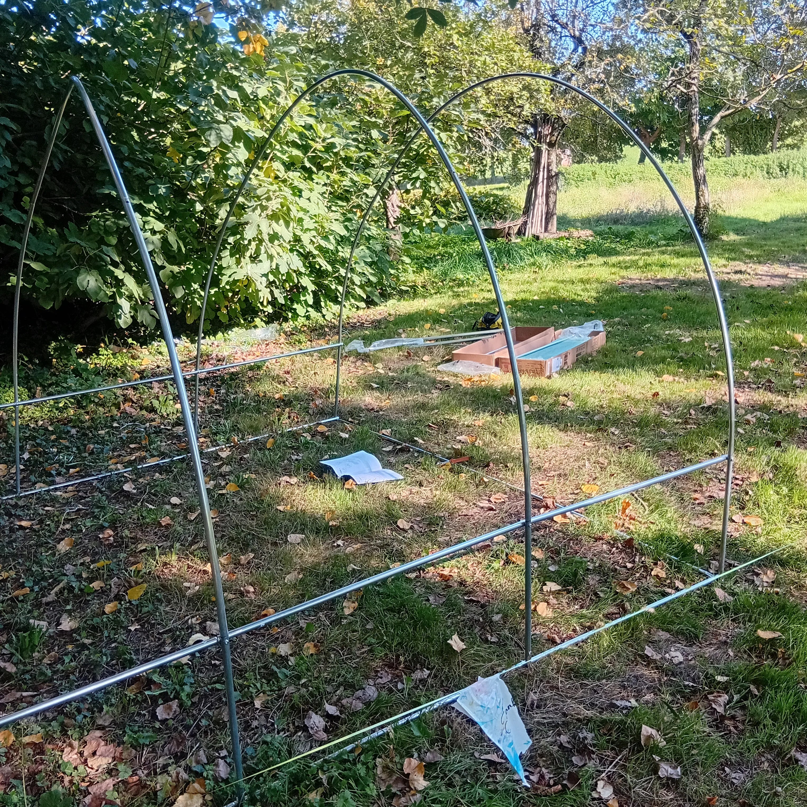 L'armature de base de la mini-serre, composée de tubes métalliques incurvés et droits, en cours d'assemblage sur la pelouse. Un manuel d'instructions est posé au sol à l'intérieur du cadre. Image