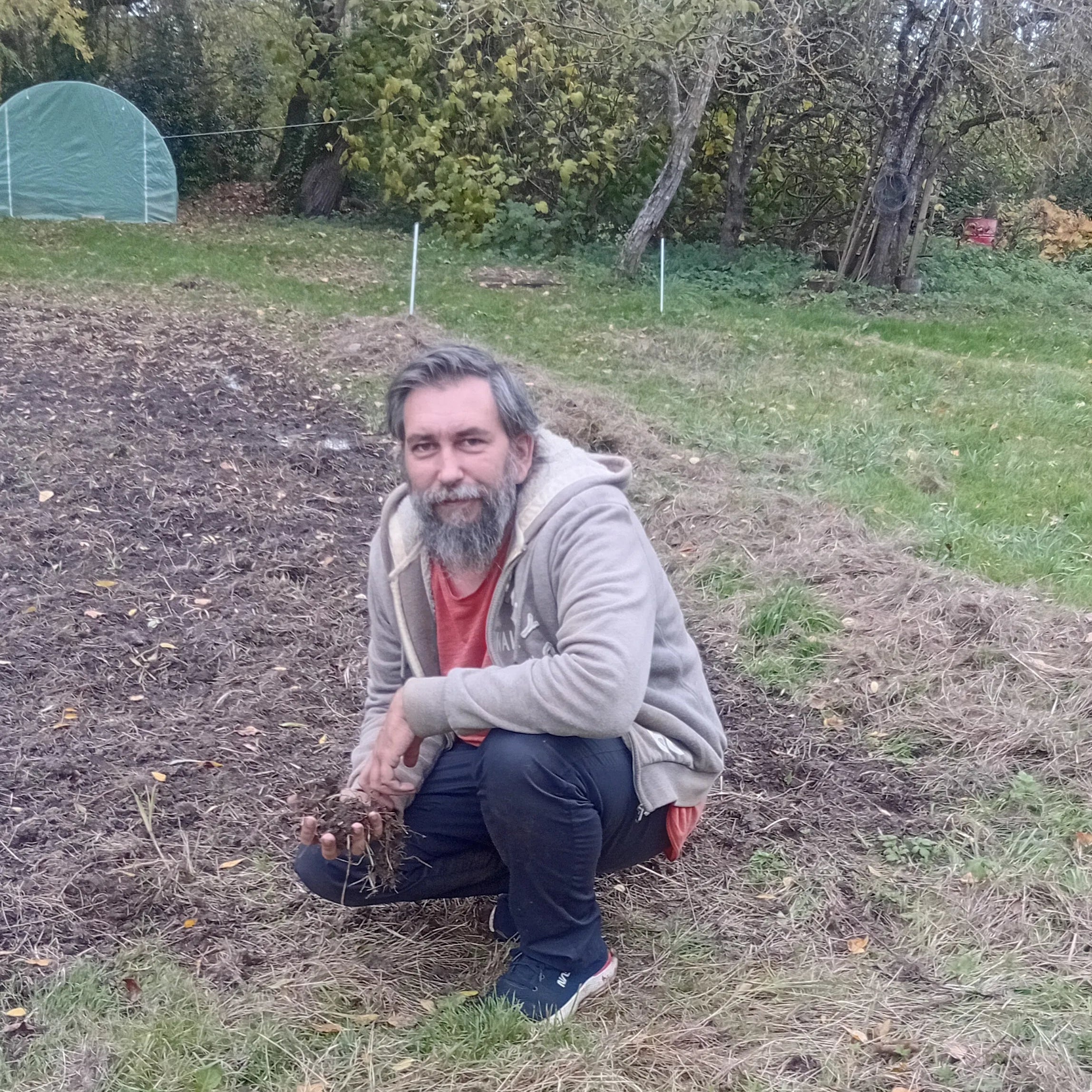Un homme d'âge moyen, avec une barbe grise, est accroupi dans un jardin en friche. Il porte un t-shirt rouge sous un gilet gris clair et un pantalon foncé. Le sol est un mélange d'herbe sèche et de terre labourée. En arrière-plan, des arbres avec des feuilles d'automne et une petite serre tunnel sont visibles sous un ciel nuageux.