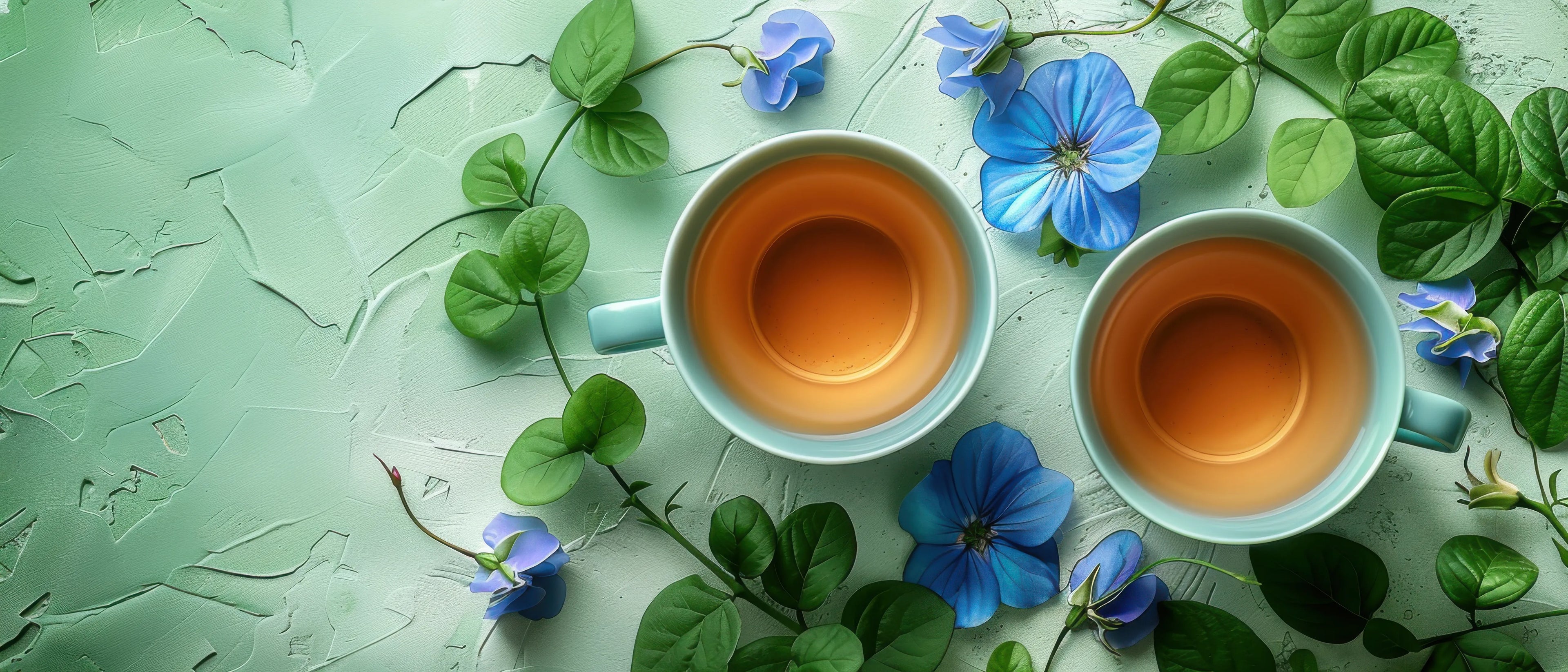 Vue aérienne de deux tasses bleu clair remplies d'un thé ambré ou doré, entourées de vignes et de feuilles d'un vert éclatant, ainsi que de magnifiques fleurs de pois papillon d'un bleu profond